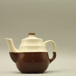 May include: A two-toned ceramic teapot with a brown base and a cream-colored upper section. The teapot features a curved spout, a rounded handle, and a lid with a small knob. The design is simple and classic, suitable for serving tea.