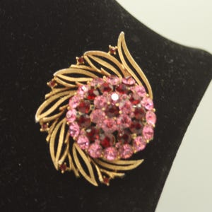 May include: A gold-toned brooch with a floral design. The center features a cluster of pink and red gemstones. The brooch has a leaf-like design around the gemstone cluster. The brooch is displayed on a black surface.