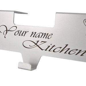 May include: A silver-coloured metal over-the-cabinet hook rack featuring a flat horizontal panel with the engraved text "Your name Kitchen" in a cursive font, flanked by floral illustrations on both ends and a single downward-facing hook.