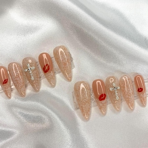 May include: Two sets of almond-shaped press-on nails. Each nail is a nude color with a lace pattern. Some nails feature a red lip design or a silver cross. The nails are displayed on clear plastic holders.