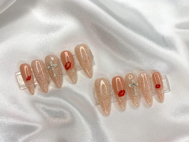 May include: Two sets of almond-shaped press-on nails. Each nail is a nude color with a lace pattern. Some nails feature a red lip design or a silver cross. The nails are displayed on clear plastic holders.