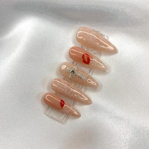 May include: A set of six almond-shaped artificial nails. The nails feature a nude base with various designs, including a red lip, a silver cross, and a lace pattern. The nails are displayed on clear plastic stands.