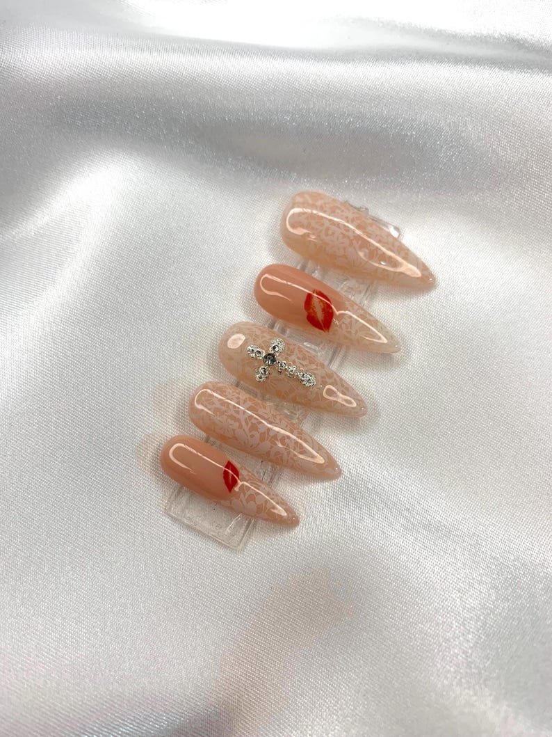 May include: A set of six almond-shaped artificial nails. The nails feature a nude base with various designs, including a red lip, a silver cross, and a lace pattern. The nails are displayed on clear plastic stands.