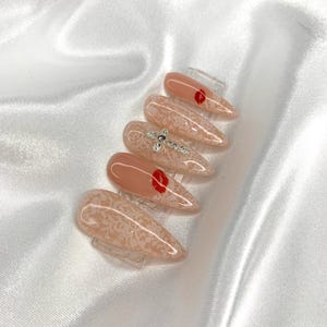 May include: A set of almond-shaped artificial nails. The nails feature a nude base with a delicate lace pattern. Some nails have a small red heart design, while others are adorned with sparkling rhinestones.