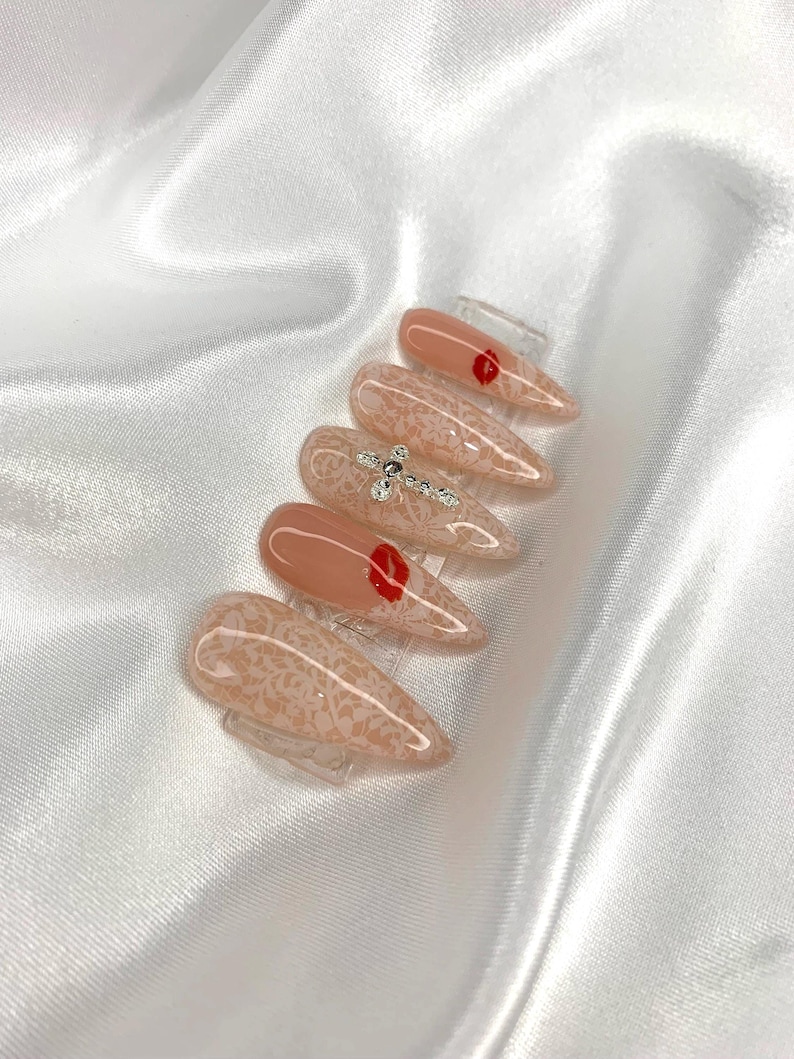 May include: A set of almond-shaped artificial nails. The nails feature a nude base with a delicate lace pattern. Some nails have a small red heart design, while others are adorned with sparkling rhinestones.