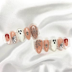 Könnte beinhalten: Zwei Sets falscher Nägel zum Thema Halloween. Jedes Set zeigt mandelförmige Nägel mit Designs wie Spinnennetz, Geist, Spinne und Blutstropfen. Die Nägel sind in Weiß, Pfirsich und Rot gehalten, mit silbernen Akzenten.