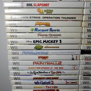 May include: A stack of 20 white Nintendo Wii game cases with colorful game covers. The games include "NHL Slapshot", "Twin Strike: Operation Thunder", "Racquet Sports", "Pirates of the Caribbean", "Epic Mickey", "Fragile Dreams", "Tornado Outbreak", "Paintball 2", "Need for Speed: The Run", "The Grim Adventures of Billy & Mandy", "Excitebots: Trick Racing", "We Cheer 2", "Target: Terror", "Petz Sports", "MySims Kingdom", "Michael Jackson: The Experience", and "Sims 3".