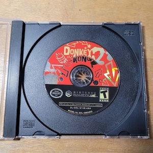 May include: A Nintendo GameCube disc for Donkey Kong 2, housed in a clear plastic case. The disc features a red and silver design with the game title and logo. The case is black with a hinged design.