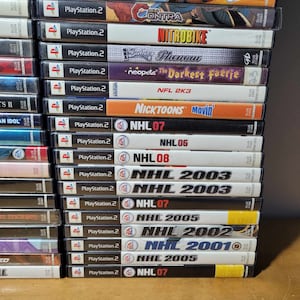May include: A stack of 20 PlayStation 2 video game cases. The cases are arranged vertically, with the spines facing the viewer. The games include titles such as "NHL 07", "NHL 2005", "NHL 2003", "NHL 2002", "NHL 2001", "Nicktoons Movin'", "NFL 2K3", "Neopets: The Darkest Faerie", "Bules Phenom", "NitroBike", "Contra", "Street", "Namco Museum 50th Anniversary", and "PlayStation 2".