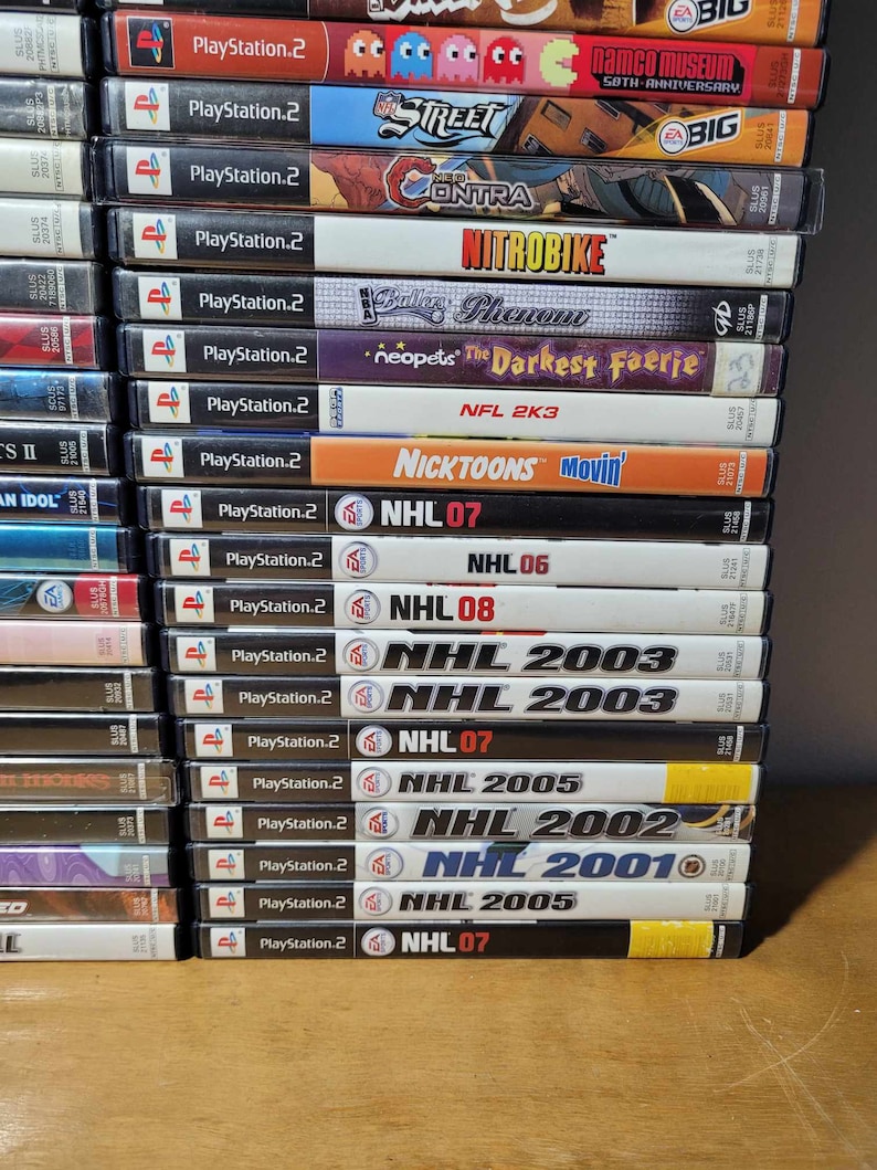 May include: A stack of 20 PlayStation 2 video game cases. The cases are arranged vertically, with the spines facing the viewer. The games include titles such as "NHL 07", "NHL 2005", "NHL 2003", "NHL 2002", "NHL 2001", "Nicktoons Movin'", "NFL 2K3", "Neopets: The Darkest Faerie", "Bules Phenom", "NitroBike", "Contra", "Street", "Namco Museum 50th Anniversary", and "PlayStation 2".