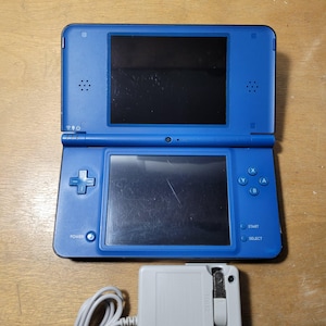 May include: A blue handheld video game console with dual screens, a directional pad, four action buttons labelled A, B, X, and Y, and Start and Select buttons. A light grey power adapter with a white cord is positioned directly below the console.