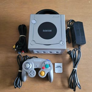 May include: Silver Nintendo GameCube console with a matching controller, power adapter, cables, and a 128MB memory card. The console has the words "NINTENDO GAMECUBE" printed on the front.