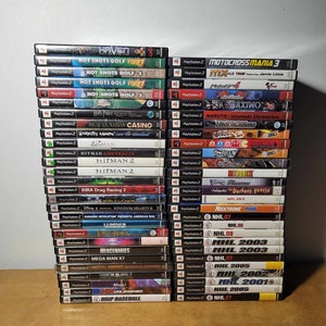 May include: A collection of 40 PlayStation 2 video game cases stacked on a wooden surface. The cases are arranged in two columns, with the titles facing forward. Some of the titles include "Haven", "Hot Shots Golf", "Motocross Mania 3", "Hitman", "NHL 2003", and "NHL 2005".