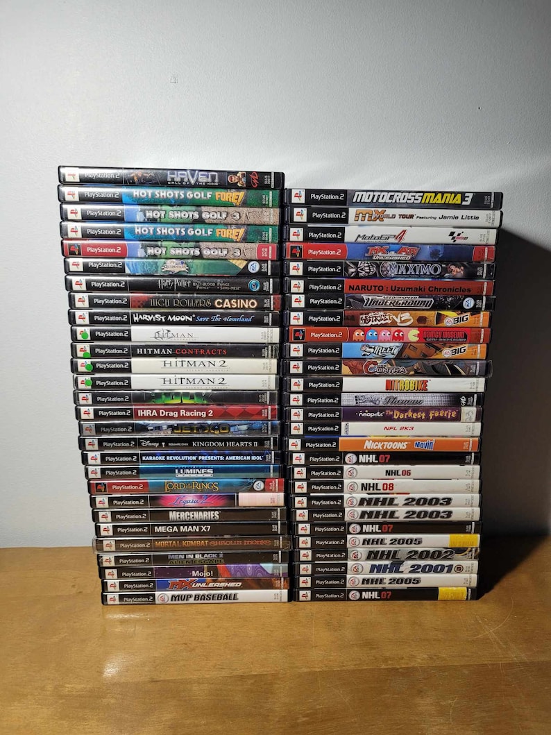 May include: A collection of 40 PlayStation 2 video game cases stacked on a wooden surface. The cases are arranged in two columns, with the titles facing forward. Some of the titles include "Haven", "Hot Shots Golf", "Motocross Mania 3", "Hitman", "NHL 2003", and "NHL 2005".