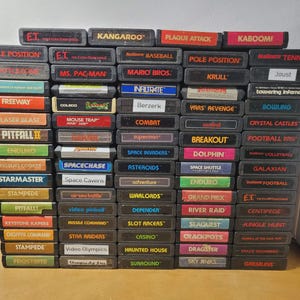 May include: A large collection of rectangular Atari 2600 video game cartridges stacked in five vertical columns. Each black plastic cartridge features a label with a unique game title printed in various colors, including E.T., Mario Bros., and Pitfall.