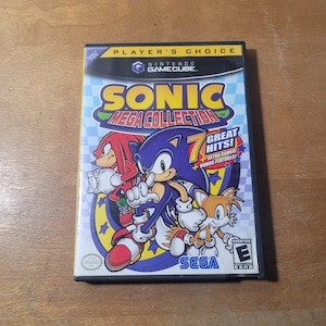 May include: A Nintendo GameCube game case for Sonic Mega Collection. The cover features Sonic the Hedgehog, Knuckles, and Tails. The game case has the text "7 Great Hits!" and "Extra Games + Bonus Features!"