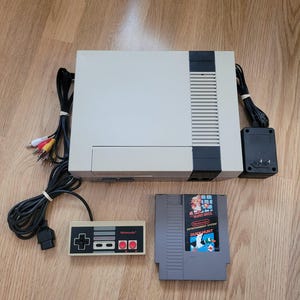 May include: A grey and black Nintendo Entertainment System console with a matching rectangular controller, a black power adapter, a multi-coloured AV cable, and a grey Super Mario Bros. and Duck Hunt game cartridge for the Nintendo system.