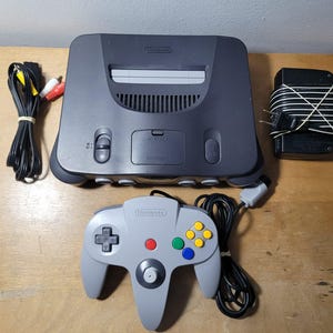 May include: A dark grey Nintendo 64 video game console with a matching light grey controller, a black power adapter with a cord, and a set of yellow, white, and red AV cables. The console features a cartridge slot, power switch, and reset button.