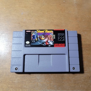 May include: A gray Super Nintendo cartridge for the game "Stunt Race FX." The label features the game's title in white and yellow, with images of race cars. The cartridge has a rectangular design with a textured surface.