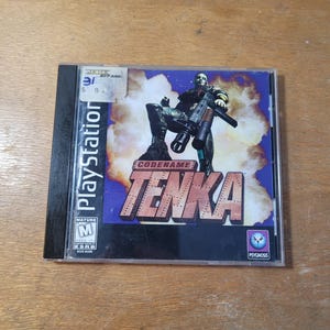 Peut inclure: Boîtier de jeu PlayStation pour "Codename Tenka", avec un soldat futuriste armé. La jaquette présente un fond bleu et orange avec le titre du jeu en grosses lettres. Le boîtier a une étiquette de prix.