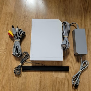 May include: A white Nintendo Wii video game console with a gray power adapter, a gray AV cable with yellow, white, and red connectors, and a black sensor bar. The console features a rectangular design with a disc slot and the Nintendo logo on top.