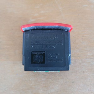 May include: A black Nintendo 64 Memory Expansion Pak with a red top. The cartridge has the Nintendo 64 logo and text that reads "MEMORY EXPANSION PAK". The model number is NUS-007 and it is made in Japan.