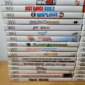 May include: A stack of 15 white Wii video game cases with colorful game titles and logos. The games include "Brothers in Arms: Double Time", "NHL 2K11", "Just Dance Kids 2", "Wipeout", "Guitar Hero III: Legends of Rock", "F1 2009", "Dora the Explorer: Dora's Crystal Kingdom", "Planet 51: The Game", "Where's Waldo? The Fantastic Journey", "Real Heroes: Firefighter", "Dance Dance Revolution Hottest Party", "The Secret Saturdays: Beasts of the 5th Sun", "Klonoa", "Star Wars: The Clone Wars: Republic Heroes", and "Yogi Bear".