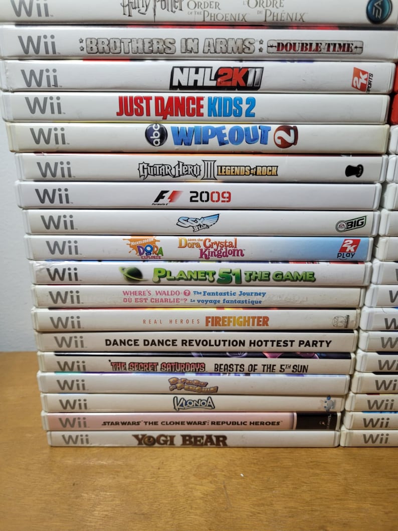 May include: A stack of 15 white Wii video game cases with colorful game titles and logos. The games include "Brothers in Arms: Double Time", "NHL 2K11", "Just Dance Kids 2", "Wipeout", "Guitar Hero III: Legends of Rock", "F1 2009", "Dora the Explorer: Dora's Crystal Kingdom", "Planet 51: The Game", "Where's Waldo? The Fantastic Journey", "Real Heroes: Firefighter", "Dance Dance Revolution Hottest Party", "The Secret Saturdays: Beasts of the 5th Sun", "Klonoa", "Star Wars: The Clone Wars: Republic Heroes", and "Yogi Bear".
