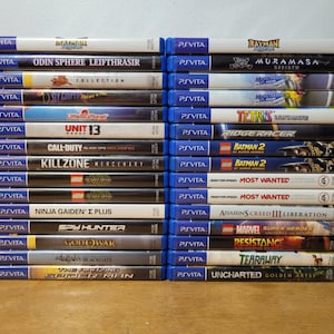 Peut inclure: Une collection de 20 cartouches de jeux vidéo pour PlayStation Vita, empilées en deux colonnes. Les cartouches sont bleues et noires avec des titres et des logos de jeux colorés. Certains des titres incluent "Odin Sphere Leifthrasir", "Muramasa", "Unit 13", "Call of Duty", "Killzone", "LEGO Batman 2", "Most Wanted", "Assassin's Creed III Liberation", "God of War", "The Amazing Spider-Man", et "Uncharted Golden Abyss".