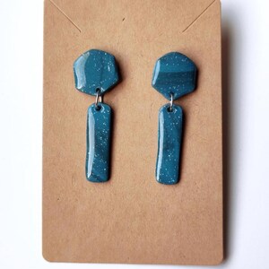 Może przedstawiać: Para kolczyków wiszących w kolorze morskim. Każdy kolczyk ma geometryczną górę i prostokątne dno, połączone srebrną pętlą. Kolczyki mają nakrapiany wzór i są prezentowane na brązowej karcie.