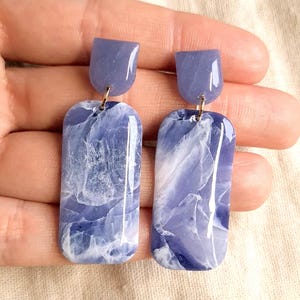 Op de afbeelding: Handgemaakte oorbellen met een gemarmerd blauw-wit ontwerp. Elke oorbel heeft een rechthoekige hanger met een glanzende afwerking, bekroond met een kleine, effen blauwe stud. De oorbellen zijn verbonden met goudkleurige metalen onderdelen.