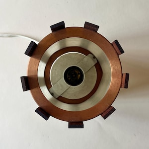 May include: Overhead view of a copper and wood lamp. The lamp has a circular design with a copper outer ring, a silver inner ring, and dark wood accents. A white cord extends from the lamp.