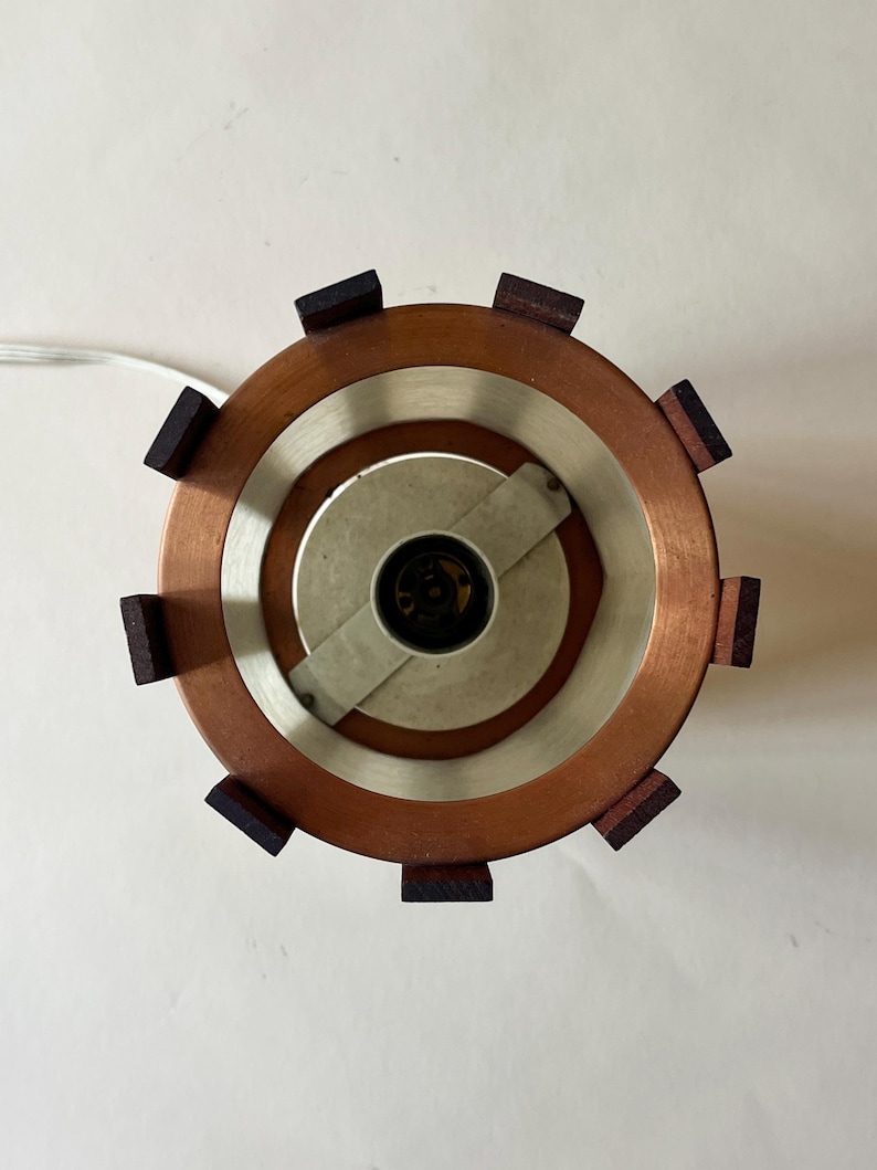 May include: Overhead view of a copper and wood lamp. The lamp has a circular design with a copper outer ring, a silver inner ring, and dark wood accents. A white cord extends from the lamp.