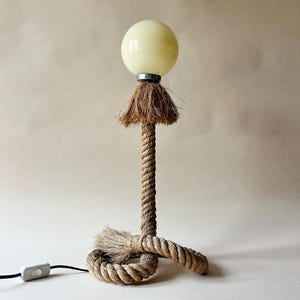 May include: A unique table lamp with a beige glass globe shade. The lamp's stem and base are made of thick, twisted rope. A small, decorative fringe sits below the globe. The lamp has a black power cord with an on/off switch.