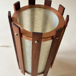 May include: A vintage cylindrical lamp with a wooden frame and a light-colored, textured shade. The lamp features vertical wooden slats held together by a circular wooden frame at the top and bottom. Small brass-colored rivets secure the slats. The lamp has a mid-century modern aesthetic.