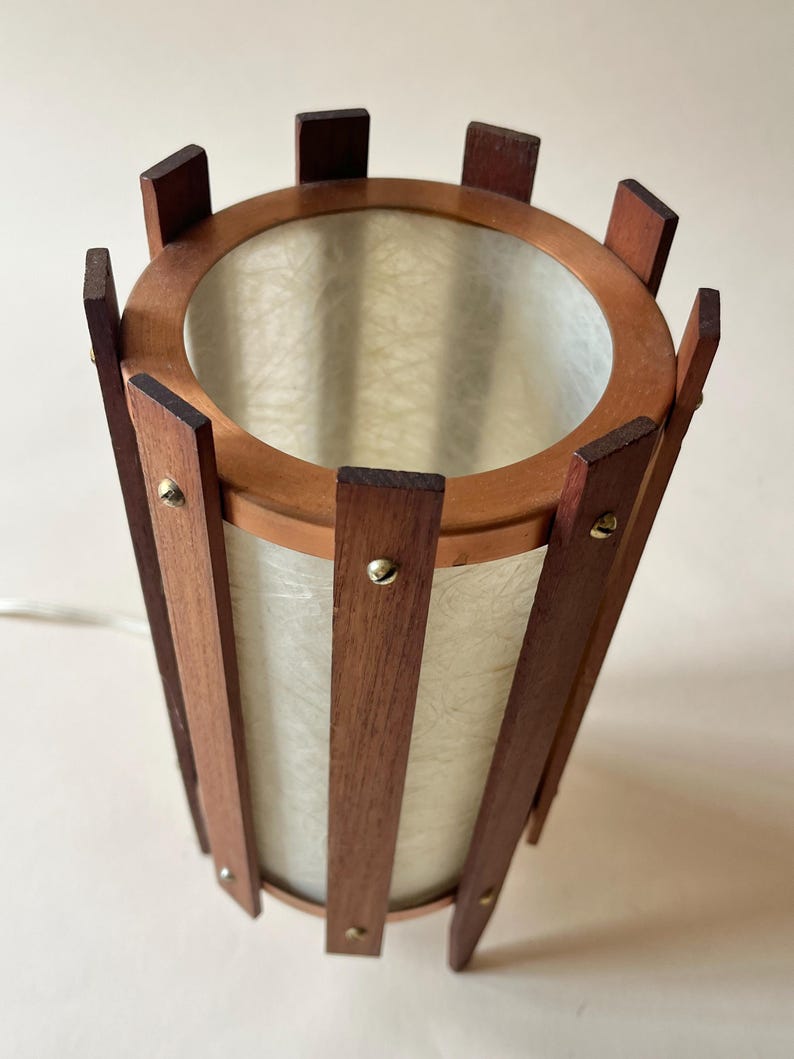 May include: A vintage cylindrical lamp with a wooden frame and a light-colored, textured shade. The lamp features vertical wooden slats held together by a circular wooden frame at the top and bottom. Small brass-colored rivets secure the slats. The lamp has a mid-century modern aesthetic.