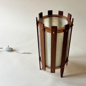 May include: A mid-century modern table lamp with a cylindrical shade. The lamp features a wooden frame with vertical slats and a circular top and bottom. The shade is a light, textured material. A white cord and switch extend from the base.