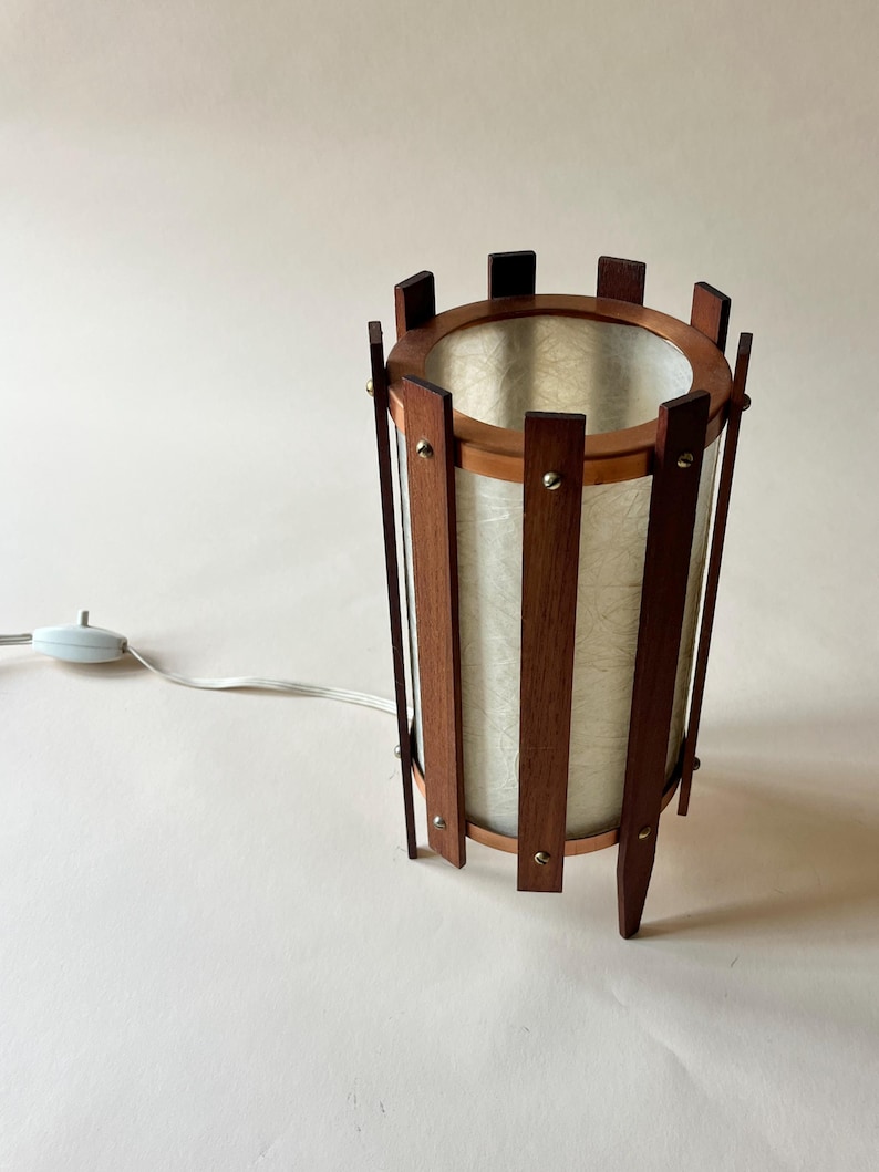 May include: A mid-century modern table lamp with a cylindrical shade. The lamp features a wooden frame with vertical slats and a circular top and bottom. The shade is a light, textured material. A white cord and switch extend from the base.