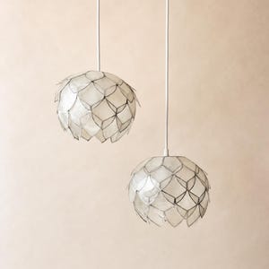 Könnte beinhalten: Zwei Pendelleuchten mit kuppelförmigen Schirmen, die aus überlappenden, durchscheinenden, cremeweißen, muschelartigen Schuppen mit dunklen, dünnen Umrandungen bestehen. Jeder Schirm hängt an einem einzelnen, dünnen, weißen Kabel.