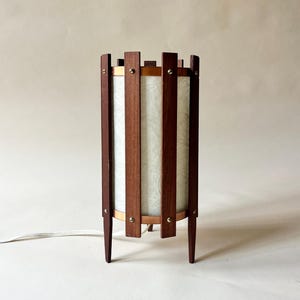May include: A mid-century modern table lamp with a cylindrical shade. The lamp features vertical wooden slats, a light-colored shade, and brass accents. The lamp has a dark wood finish and stands on four tapered legs.