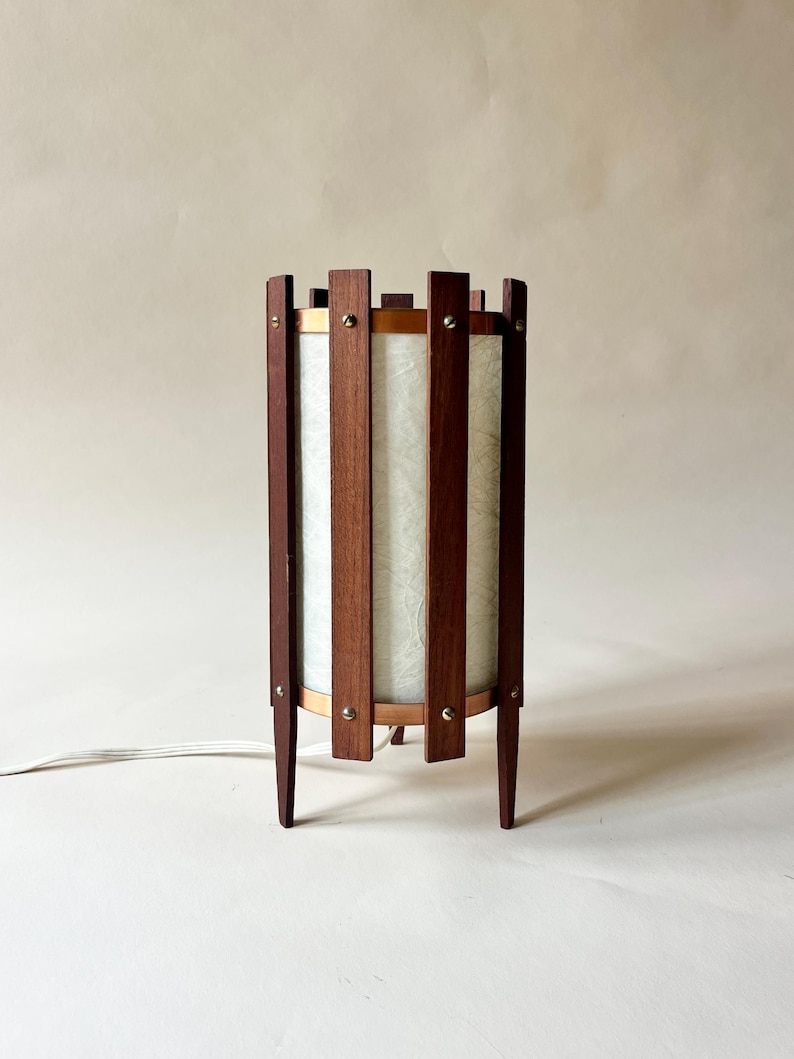 May include: A mid-century modern table lamp with a cylindrical shade. The lamp features vertical wooden slats, a light-colored shade, and brass accents. The lamp has a dark wood finish and stands on four tapered legs.