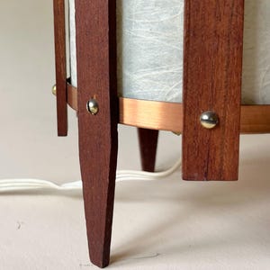 May include: A mid-century modern lamp with a cylindrical shade and a wooden frame. The frame is made of dark brown wood and features a copper band. The lamp has three legs and a white cord.