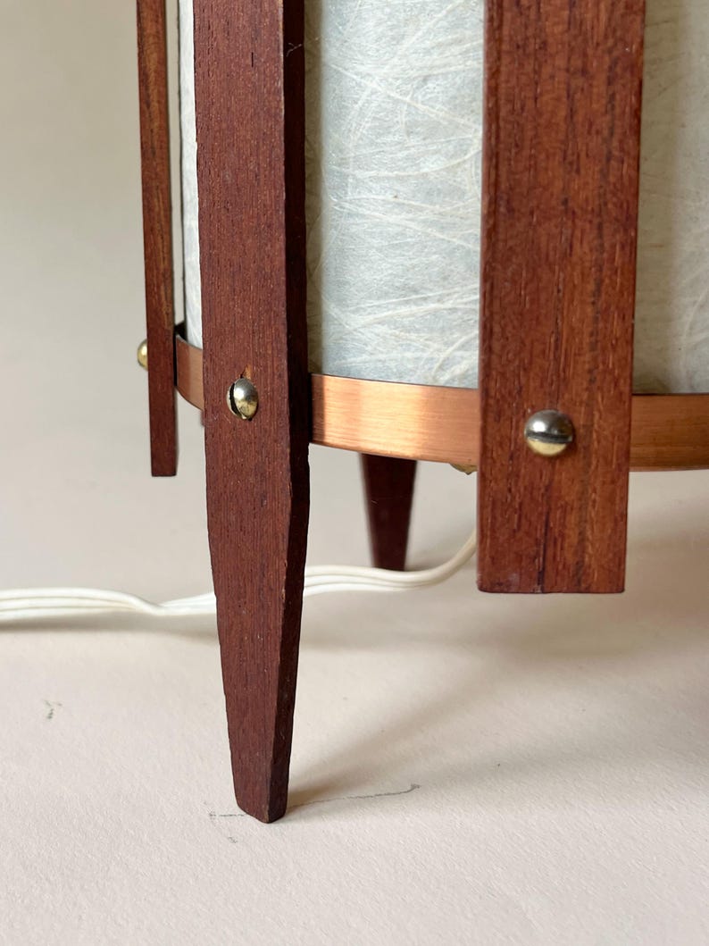 May include: A mid-century modern lamp with a cylindrical shade and a wooden frame. The frame is made of dark brown wood and features a copper band. The lamp has three legs and a white cord.