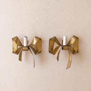 Op de afbeelding: Een paar wandlampen van goudkleurig metaal, elk gevormd als een decoratieve strik. Elke wandlamp is voorzien van een centrale witte cilindrische kaarsvormige fitting die in het midden van de metalen strikstructuur is geplaatst.