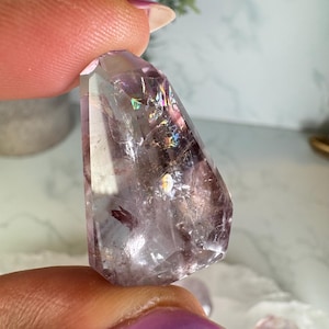 Puede incluir: Un cristal facetado y translúcido con un tono púrpura claro. El cristal tiene iridiscencia interna similar al arcoíris y se sostiene entre dos dedos. La superficie del cristal tiene un aspecto pulido y los bordes están claramente definidos.