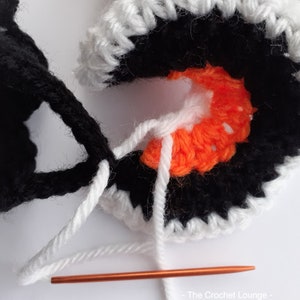 May include: Close-up of a crochet project in progress. The design features black, white, and orange yarn, with a copper crochet hook. The project appears to be a circular motif, with the white yarn being used to connect the pieces.