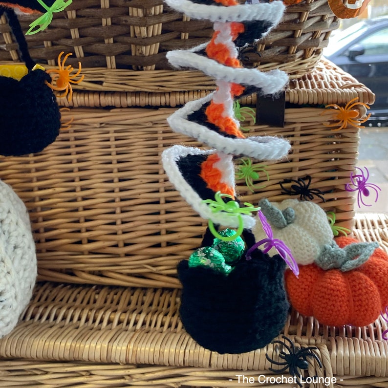 May include: A collection of handmade crochet Halloween decorations. Includes a black cauldron with green candies, a white and orange spiral, a pumpkin, and spiders. The decorations are set against a wicker basket.