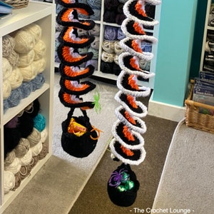 May include: Two crochet Halloween decorations. One is black, orange, and purple, with a small black bag holding candy. The other is white, orange, and black, with a small black bag holding green and purple candies. The Crochet Lounge is in the bottom right corner.