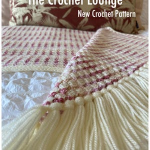 May include: A cream-coloured crocheted blanket with pink accents and fringe. The blanket is folded over a white surface. The text "The Crochet Lounge New Crochet Pattern" is visible in the image.