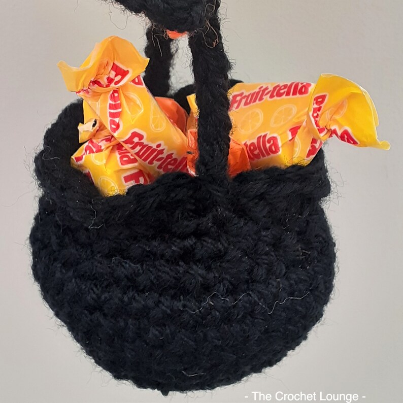 May include: A small, black crocheted basket filled with orange and yellow wrapped candies. The basket has a rounded shape with a handle. The candies are labeled "Fruit-tella". The Crochet Lounge is printed at the bottom right.